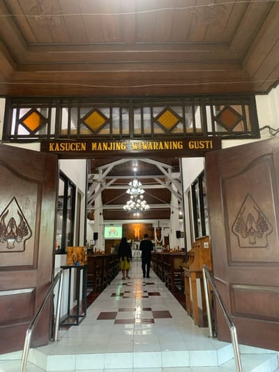 The image shows the entrance to a church in Kecamatan Kartasura, Indonesia (latitude: -7.567, longitude: 110.757). The entrance is framed by two wooden doors with carved designs. Above the entrance, there is a sign that reads "KASUCEN MANJING WIWARANING GUSTI." A stained glass window is visible above the sign. The interior of the church is visible through the open doorway, with rows of wooden pews and a chandelier hanging from the ceiling. There are two people walking towards the altar. The floor is tiled in a checkered pattern.
