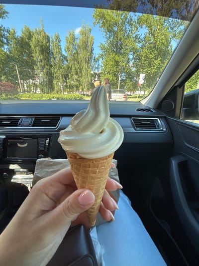 In the city of Fedorino, Russia, a person is holding a soft-serve ice cream cone inside a car. The ice cream is vanilla-flavored and presented in a waffle cone. The car's interior, including the dashboard with air vents and a car stereo, is visible. Outside the car, through the windshield, a sunlit scene with green trees and blue sky can be observed. Two individuals are visible in the background, standing near a car. The overall setting appears to be a warm, sunny day.