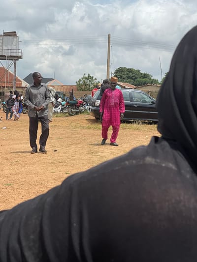 The image captures an outdoor scene in Kassa, Nigeria, under a cloudy sky. In the foreground, a person wearing a black hijab is partially visible, facing away from the camera. Two men are the main subjects in the midground. The man on the left is wearing a grey patterned shirt and dark pants, looking towards the left of the frame. The man on the right is dressed in a vibrant pink traditional outfit and a matching cap, with his gaze directed forward. Behind them, a black sedan is parked, and several motorcycles are visible to the left of the car. In the background, buildings with corrugated iron roofs and a water tower are present, along with trees and a utility pole. The ground is covered in loose, sandy soil. The overall atmosphere appears to be that of a public gathering or event.