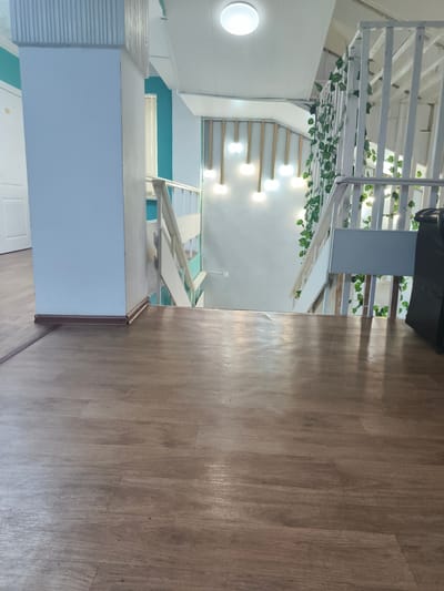 This is a photograph taken at a downward angle, looking across a wooden floor towards a staircase. The floor is made of light brown planks with a visible wood grain pattern. To the left, a large light blue pillar is prominent, with a ribbed silver section at the top and a white door visible behind it. The pillar has a dark brown baseboard. The staircase, made of white painted metal and wood, curves to the right and descends out of frame. Green artificial vines are draped along the white railings of the staircase. The walls in the background are painted in a light blue and teal color scheme. Several bright, circular light fixtures are visible on the ceiling. The environment appears to be an indoor common area, potentially a hallway or lobby leading to a stairwell. The location is identified as Arkalyk, Kazakhstan.