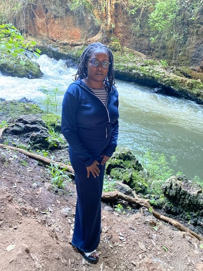 A medium shot captures a woman standing in what appears to be a cave or grotto-like area. The location seems to be outdoors, with a stream or river flowing in the background and lush greenery surrounding the scene. The woman is positioned towards the right side of the image frame, and she is wearing glasses and has her hair in dreadlocks. She is clad in a navy blue tracksuit with a possible striped shirt visible at the neck.
Her hands are gently placed on her stomach. The background reveals a rugged terrain with exposed rocks, moss, and vegetation. The water appears to be flowing with some force, suggesting a natural water feature like a waterfall or a rapid stream. The environment is characterized by the natural elements of a cave setting, with a mix of light and shadow playing across the rocks and foliage. This scene takes place in or around Kianyaga, Kenya.