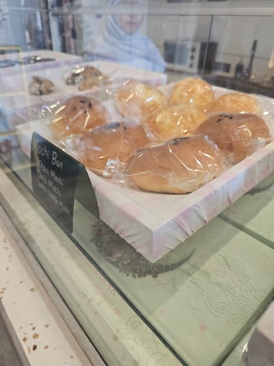 In a display case at a bakery in Banyuurip, Indonesia, an assortment of individually packaged mochi buns are presented. The buns are golden brown and some appear to have dark speckles, possibly chocolate chips or sesame seeds. A black sign in the foreground reads "Mochi Bun," followed by "Aka Mochi," "Choco Mochi," and "Sweet Cheese Mochi." The buns are arranged on a white and pink checkered cloth. In the background, a person wearing a light-colored hijab is partially visible, and behind them, a blurred view of a kitchen or serving area can be seen. The display case reflects the surrounding environment, including what appears to be patterned boxes with the text "HIKMAT MODAIKAN" and "GRACEFUL AND JOYFUL EID." The overall scene depicts a retail setting focused on baked goods.