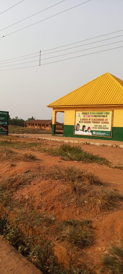The image displays an outdoor scene in Ketu Adiowe, Nigeria, featuring a recently constructed or renovated school building under a clear daytime sky. The building in the foreground on the right has a distinctive bright yellow corrugated metal roof and a two-toned facade with a yellow upper section and a dark green lower band.

A large banner is prominently displayed on the building, stating its sponsorship: "SPONSORED BY: THE FEDERAL GOVERNMENT OF NIGERIA." The banner's project title reads: "CONSTRUCTION OF A BLOCK OF CLASSROOMS AT KETU ADIOWE PRIMARY SCHOOL, ADO-ODO OTA LOCAL GOVERNMENT." It also features photographic portraits of three men, likely public figures or officials. To the left, another partially visible sign further indicates "GOVERNMENT OF NIGERIA" and mentions a "MINISTRY OF... TECHNOLOGY."

In the middle ground, behind the main structure, several other low-rise buildings with brown roofs are visible, suggesting an extended school facility. The ground in the foreground and around the buildings is characterized by reddish-brown dirt, with sparse patches of dry green grass and small plants. Overhead, multiple thin power lines stretch across the pale, light blue sky, with one vertical support pole visible among them. The lighting is soft and even, indicative of a clear day, possibly morning or late afternoon. The scene portrays a public infrastructure development in a rural or semi-urban Nigerian setting.