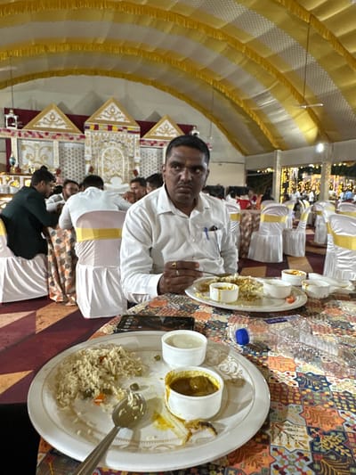 Captured indoors, this scene features a seated man in Nashik, India. He is dressed in a white shirt and is positioned at a table laden with food. The table, covered in a patterned cloth, displays plates of rice and containers of curry. Other individuals are also seated at tables, creating a sense of a shared dining experience. The environment includes a decorated archway and ornate decorations, suggesting a formal or celebratory event. The photo is taken at an angle, providing a broader view of the setting.