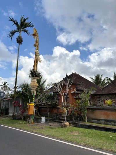 The image shows a street view in Susut, Indonesia. On the right side of the frame is a traditional Balinese building with a red tiled roof and intricate carvings on its stone walls. In front of the building, there is a decorative pole adorned with woven offerings and what appears to be a palm frond. To the left of the decorative pole, there is a tall palm tree. A road with a white line marking the lane runs along the bottom of the image. The sky is blue with scattered white clouds. The surrounding vegetation is lush, with more palm trees and green grass. There are no people visible in the image.