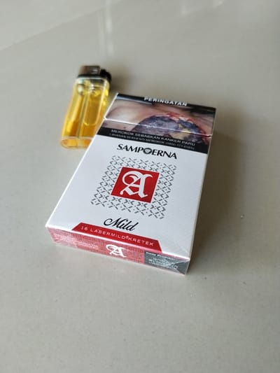 A close-up, eye-level shot shows a cigarette pack and a lighter on a light-colored, textured surface, possibly a table or floor. The cigarette pack is white with red accents and features the "Sampoerna A" logo. Text on the pack indicates "Mild," "16 LASERMILD KRETEK," and a warning about smoking causing lung cancer. The lighter is clear with yellow fluid and a silver top. The background is blurred, suggesting a shallow depth of field. There are no people present in the image. The scene appears to be set indoors, given the lighting and the surface material. There is no information available regarding the specific location within Panteepanah, Indonesia.