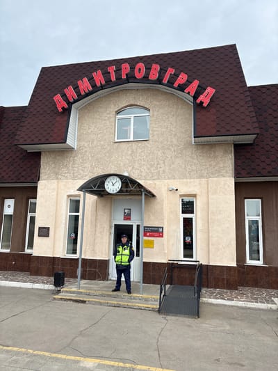 The scene depicts the exterior front of a public building in Dimitrovgrad, Russia. The building is identified by large red Cyrillic letters spelling "ДИМИТРОВГРАД" mounted on its dark brown shingled roofline. The facade consists of a light beige textured stucco finish above a brown tiled lower section, featuring several white-framed windows. A central entrance is covered by a dark-framed canopy from which a circular white clock, displaying approximately 12:20, is suspended.
A male individual, appearing to be a security guard or official, stands centrally in the doorway, facing forward. He wears a dark blue uniform comprising a cap, jacket, and trousers, overlaid with a high-visibility lime green vest featuring reflective stripes. To the right of the entrance, a black metal wheelchair ramp with handrails extends from the tiled sidewalk to the asphalt ground level. To the left of the entrance, on the sidewalk, a black speaker on a stand is positioned near the steps. The ground in the foreground is cracked asphalt pavement, marked with a yellow line, leading to a patterned tiled sidewalk directly in front of the building. The sky overhead is uniformly overcast.