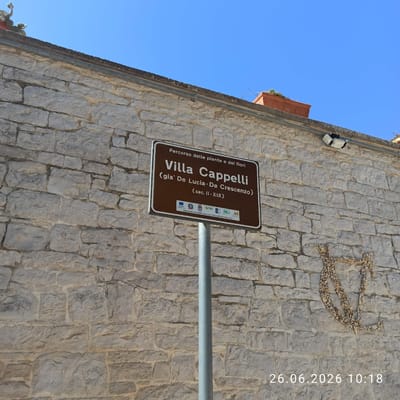 This media file is a photograph of a signpost in front of a stone wall. The sign is brown with white text and is tilted at an angle. The text on the sign reads: "Percorso delle piante e dei fiori Villa Cappelli (gia' De Lucia - De Crescenzo) (sec. II-XIX) OPERAZIONE COFINANZIATA DAL P.S.R. PUGLIA 2007-2013 MIS, 313 AZ 3". There are also several small logos visible on the lower right side of the sign. Above and to the left of the sign, a bright blue sky is visible. The stone wall behind the sign appears to be made of irregular, light-colored stones. There is also a decorative element made of small stones forming a pattern on the wall. A metal pipe runs horizontally across the image just above the signpost. A timestamp "26.06.2026 10:18" is overlaid on the image on the right side. The image appears to be taken outdoors during the day, with clear skies. The overall impression is that of a historical or informational sign in an outdoor setting, possibly a park or garden area given the mention of "piante e dei fiori" (plants and flowers).