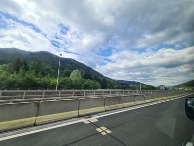 The image captures a highway view in Steinhaus am Semmering, Austria, characterized by a concrete barrier and a metal railing separating the road from a tree-covered mountainside under a cloudy sky. The road surface is dark asphalt with white lane markings. In the distance, a hill dotted with trees slopes upwards, and several streetlights are visible along the highway. The sky is filled with a mix of gray and white clouds, with patches of blue peeking through, suggesting dynamic weather conditions. The reflection in the side mirror of a vehicle shows a similar cloudy sky. No people are visible in the frame.