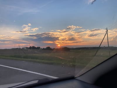 The image depicts a sunset over a rural landscape. The sun is partially obscured by clouds, casting a warm orange glow across the sky. The clouds are scattered, with some illuminated by the setting sun and others in shadow. A line of trees is visible on the horizon, silhouetted against the colorful sky. In the foreground, a road with a white line runs diagonally across the frame, suggesting the perspective is from inside a vehicle. To the right, a power pole with wires stretches upwards, also silhouetted. The fields appear to be agricultural, with some areas of green and others looking dry or harvested. The overall impression is one of a peaceful evening in a rural setting, likely in or near Самахвалавіцкі сельскі Савет, Belarus, given the characteristic landscape and power infrastructure.