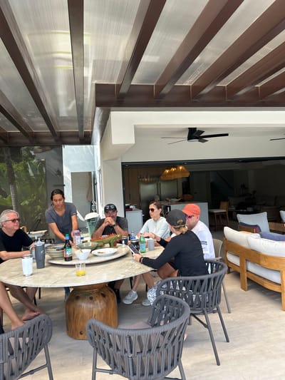 The image depicts a group of people sitting around a large, round marble table on a patio with a brown pergola roof, located at a resort in Playa Herradura, Costa Rica. The patio floor is made of light-colored tiles. The group is composed of six individuals. Two women and two men are seated around the table. There is a woman standing behind the table to the left. One man is seated off-camera to the right. The man to the left of the table is wearing a blue shirt and dark pants. The woman to his right is wearing a dark blue shirt and dark pants. The man seated to the right of the table is wearing a black shirt and dark pants. The woman to his left is wearing a black shirt and grey pants. The woman to her right is wearing a red and white shirt. The man seated to her right is wearing a blue shirt and black pants. There is a plastic iguana figurine on the table. The woman standing behind the table is wearing a grey shirt and dark pants. All the people are looking down at the table. The table is surrounded by wicker chairs. There is a wicker couch and two chairs located to the right of the table. There is a stainless steel refrigerator and a white sink visible in the background through a large glass door. There is a ceiling fan on the ceiling. The table is covered in a white tablecloth and a variety of items, including a glass of orange juice, bottles of water, a plate of food, a plate of fruit, and a plate of silverware. The overall mood of the image is one of casual relaxation.