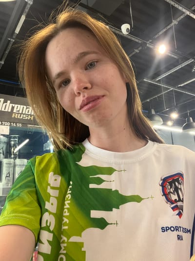 A woman is posing for a selfie. She has light brown hair and blue eyes. She is wearing a white t-shirt with a green silhouette of a Russian Orthodox church. The t-shirt also has a patch with the text "SPORT RISM RIA" and the year 1895. Behind her, there is a shelf with cans of "Adrenaline Rush" energy drinks. Text on the energy drink reads "Adrenaline zero sugar". In the background, there is a metal ceiling structure with overhead lights. The text on the shirt says "ОМУ ТУРИЗМ" which translates to "to tourism" in English. The shirt design suggests she is in Казань (Kazan), Russia, with the depicted churches representing the Kazan Kremlin.