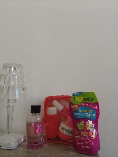 An indoor, eye-level, medium shot captures a still life of cosmetic products and a lamp, likely on a shelf or countertop. The background is a plain, light-colored wall. In the foreground, to the left, sits a clear, faceted glass lamp with a white base. To the right of the lamp, a clear glass perfume bottle with a black cap contains a pink liquid and a pink label with a black "K" logo. Next to it, a clear plastic bag holds several red-colored cosmetic containers, including a cylindrical bottle with a white cap and a small jar with a white lid. To the far right is a bright pink, pouch-like container with colorful graphics and text that appears to be a supplement or cosmetic product. The lighting is soft, casting faint shadows, suggesting an indoor setting with ambient light. The scene is devoid of any human presence or discernible activity. The location of Nor Geghi, Armenia, is not indicated by any visual elements in the image.