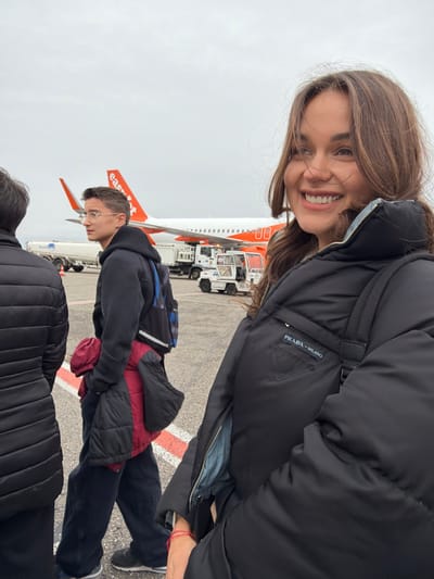The image depicts an outdoor scene on an airport tarmac at Lyon-Saint Exupéry Airport in Colombier-Saugnieu, France, under an overcast sky. In the foreground on the right, a young woman with long brown hair smiles, looking towards her right, off-camera. She wears a black puffer jacket featuring a "PRADA - MILANO" triangular logo patch on the left chest and a red string bracelet on her right wrist. Behind her, in the mid-ground, a young man with short brown hair and eyeglasses wears a dark hooded sweatshirt and dark trousers, carrying a dark backpack and a red and black puffer jacket over his left arm. He is looking to his left. Partially visible on the far left foreground is the back of a third individual wearing a dark puffer jacket. In the background, an EasyJet aircraft, white with an orange tail and "easyJet" logo, is parked on the tarmac. Various white ground support vehicles, including a fuel tanker truck with "World Fuel Services" visible, are also present on the tarmac. Red and white markings are visible on the tarmac surface. The individuals appear to be walking in an open-air airport environment.