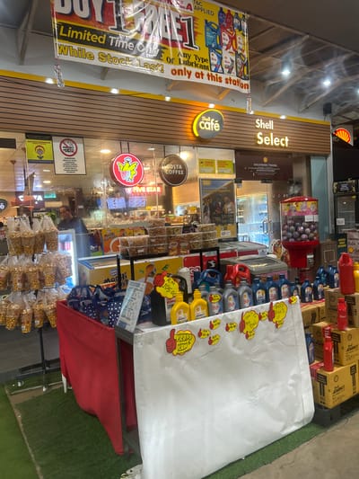 The image depicts the interior of a Shell Select convenience store and a Shell petrol station in Gurun, Malaysia. In the foreground, a promotional display is set up with various engine oil bottles, priced with red tags. A large white sheet with handwritten promotional information and yellow "thumbs up" stickers is draped over a table. Bags of snacks are displayed on shelves to the left, and a red cloth covers a section of the display table. Behind the promotional area, a man is visible inside the store, near a cooler. The Shell Select signage is prominently displayed above the store entrance. The store appears to be part of a larger petrol station complex, as indicated by the Shell branding. The overall environment suggests a retail setting within a petrol station.