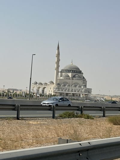 A large, white mosque with a tall minaret and multiple domes is visible on the right side of the frame. The mosque appears to be made of stone or concrete and has intricate architectural details. In front of the mosque, there is a highway with several cars driving on it. The highway is bordered by a concrete barrier and dry, scrubby vegetation. The sky is clear and pale blue, suggesting a sunny day. The overall impression is of a modern city with religious and infrastructural elements coexisting. Based on the distinctive architecture, this is likely the Al Noor Mosque in Sharjah, United Arab Emirates.