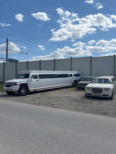 In Komsomol's'ke, Ukraine, a fleet of limousines is parked on a gravel lot adjacent to an asphalt road. The sky above is a vivid blue with scattered white clouds, and a tall, gray, paneled wall runs along the background, possibly a sound barrier.

Dominating the left side of the frame is a white Hummer limousine, its extended body stretching impressively long. Its chrome accents gleam under the sunlight, and the vehicle sits on rugged tires. To its right, a light green, older model sedan is parked, with its license plate visible. Further to the right, a white Chrysler 300 limousine is positioned, its distinctive grille prominent. All vehicles appear to be stationary, with no occupants visible. The ground is covered in loose gravel, with patches of dry grass. Overhead, power lines and a lamppost are present, suggesting an urban or semi-urban setting.