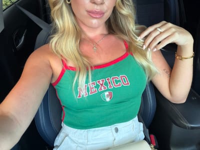 A woman with blonde hair is sitting in the driver's seat of a car. She is wearing a green tank top with red trim that says "MEXICO" in red letters and has the Mexican flag emblem. She is also wearing white shorts. The woman is wearing a gold necklace with a cross pendant, a gold ring on her left hand, and a gold bracelet on her right wrist. She has a tattoo on her right arm. The car appears to be parked in an outdoor setting, possibly on a street or in a parking lot in Pompano Beach, Florida.