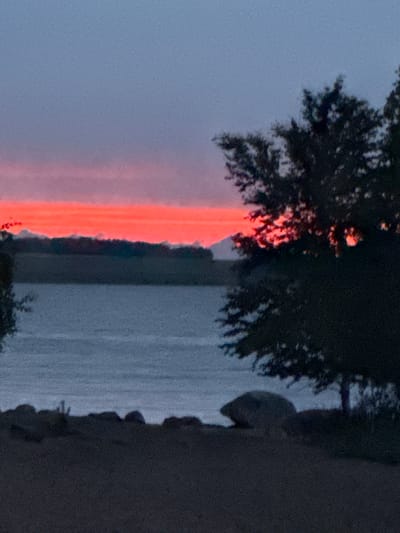 This media file depicts an outdoor scene with a prominent, vibrant orange streak of light, likely a sunset or sunrise, cutting vertically through the frame. This streak illuminates a section of dark foliage, possibly a tree, that frames the left side of the image. Beyond the foliage and the fiery light, a body of water, likely a lake or river, stretches into the distance, its surface appearing calm and reflecting the muted sky. The right side of the image is dominated by a dark, solid mass, possibly a rock formation or a man-made structure, which creates a strong contrast with the natural elements on the left. The overall mood is serene and tranquil, with the dramatic color of the light creating a focal point. The time of day is clearly either dawn or dusk. There is no visible text in the image.