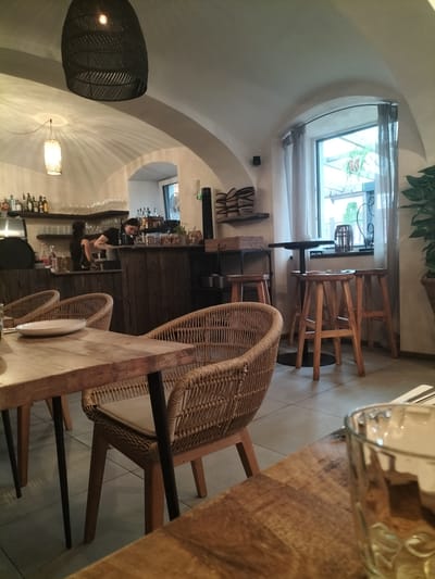 The image depicts the interior of a restaurant in Pardubice, Czechia. In the foreground, a wooden table is set with a white plate and cutlery. A woven chair with a beige cushion is positioned at the table. The restaurant's decor features vaulted ceilings, warm lighting from pendant lamps, and wooden furnishings. In the background, a bar area is visible with shelves stocked with bottles and glassware. A person is seen behind the bar, appearing to be working. A large window with sheer curtains allows natural light to enter the space, and outside greenery is visible. Bar stools and a high table are also present near the window, suggesting a separate seating area. The overall atmosphere appears to be cozy and inviting.