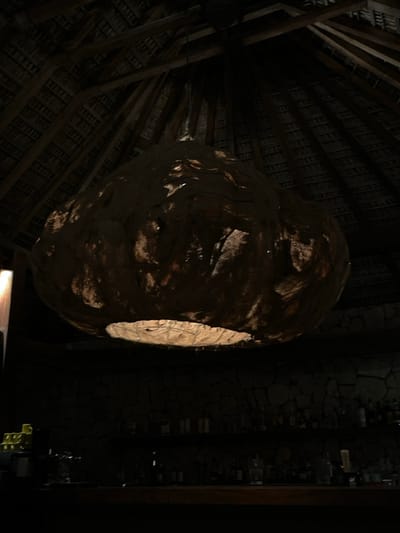The image shows a large, round, dark, wooden lamp hanging from the ceiling of a bar in San Isidro Llano Grande, Mexico. The lamp is lit from the inside, casting a warm glow. The ceiling is made of wood and the walls are made of stone. There are shelves behind the bar containing many bottles of liquor. The bar is empty, with no people present. The scene is dark and dimly lit. The coordinates provided are: latitude: 15.939, longitude: -97.274.