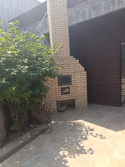 The image shows an outdoor area with a brick barbecue structure and a large bush. The barbecue is made of light-colored bricks and has two openings, one above the other. A dark brown slatted wall is visible to the right of the barbecue. The ground is paved with large light-colored tiles, and shadows from leaves are cast on the ground, indicating sunlight. A coiled hose is lying on the ground near the bush. The area appears to be a backyard or patio space. Based on the provided information, this scene is located in Buzovna, Azerbaijan. There are no people or animals visible in the image.