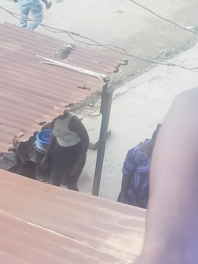 The image shows a scene in Alalubosa, Nigeria. A person wearing a dark patterned shirt is partially visible near a section of corrugated metal. A black and white goat is also visible, standing near a blue bucket. The background appears to be a concrete or stone surface.