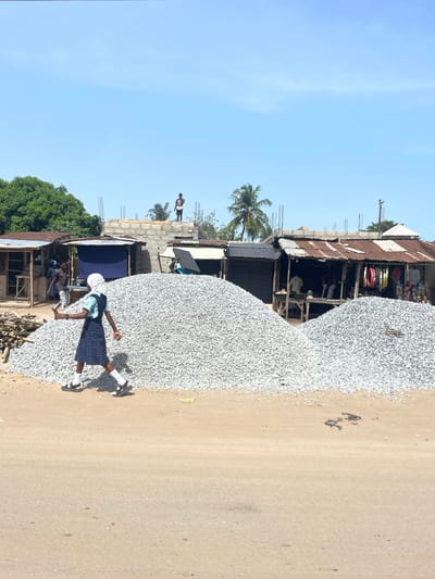 A street scene in Ikoga, Nigeria, shows a large pile of gravel alongside a dirt road. A person in a school uniform is walking beside the gravel pile. A row of shacks line the other side of the road, with people visible in and around them. A concrete building under construction is visible in the background. A palm tree is seen behind the shacks. The sky is clear and blue.