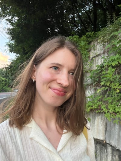 A young woman with light brown hair and blue eyes is pictured from the chest up, smiling slightly. She is wearing a cream-colored collared shirt and a small hoop earring in her left ear. The background consists of lush green foliage growing on a concrete wall and trees, with a road and some vehicles visible in the distance on the left. The setting appears to be a natural or semi-natural area in Pocheon-si, South Korea.