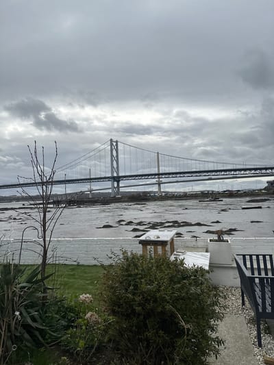 The image depicts a panoramic view of the Firth of Forth in North Queensferry, United Kingdom, under an overcast, grey sky. In the mid-ground, three bridges are visible: the prominent Forth Road Bridge (suspension bridge) with vehicle traffic on its deck, the Queensferry Crossing (cable-stayed bridge) partially visible behind it to the right, and the Forth Bridge (cantilever railway bridge) in the far background on the left.

The foreground features a garden area. On the left, a bare deciduous tree with developing buds stands prominently, partially obscuring the bridges. Below it, green grass and various plants, including some with dried, light-brown flower heads, are present. On the right, a low, white-painted brick wall runs horizontally. Behind this wall, a light-colored outdoor table with a stone or marble top and wooden legs holds two small, smooth stones. A second, similar light slab is to its right. A white square planter contains a small plant. A dense green bush occupies the immediate right foreground. Further right, a dark-colored metal slatted bench rests on light gravel, which transitions to a concrete path and then to grass. The water of the Firth of Forth appears calm, revealing extensive muddy or silty foreshore with patches of dark seaweed or vegetation, indicative of low tide. Several small boats are moored on the distant opposite bank. No people are visible.