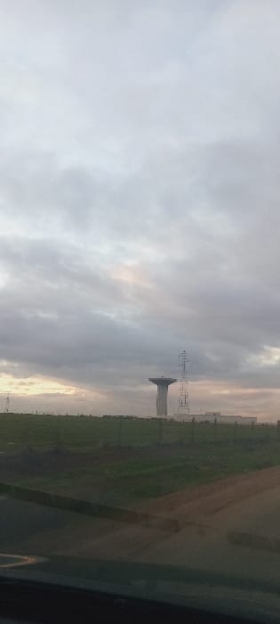 An overcast sky dominates the image. Below, a green field transitions to an earthy tone further back. A tower structure is visible in the middle ground, accompanied by utility poles stretching across the horizon. A dark form appears in the foreground, likely a window frame or vehicle interior. The scene is bathed in soft, muted light due to the heavy cloud cover.