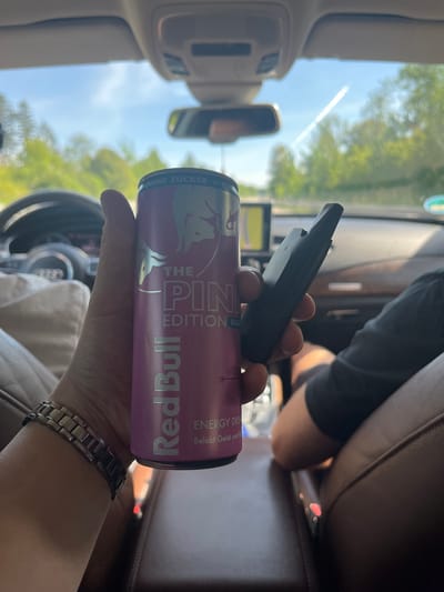 A person's hand, wearing a metal watch, holds a pink can of Red Bull "The Pink Edition" and a black vape pen inside a car. The car interior is visible, with brown leather seats and a dashboard. Through the windshield, trees and a blue sky with a contrail are visible, suggesting an outdoor, possibly rural, setting. The location is identified as Ohlstadt, Germany.