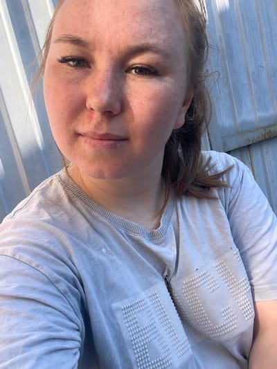 A young woman with fair skin and auburn hair pulled back in a ponytail is looking directly at the camera. She has freckles across her nose and cheeks and appears to be wearing light makeup, including eyeliner and mascara. A small red mark is visible on her upper lip. She is wearing a light gray t-shirt with a textured pattern on the chest, resembling raised dots forming letters. The background is a corrugated metal fence, likely a common sight in the industrial or utilitarian areas of a city like Aryazh, Russia. The lighting suggests it might be late afternoon or early evening, with the sun casting a warm glow on her face.