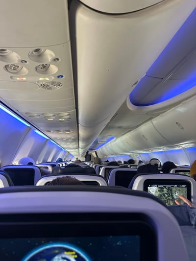 The airplane cabin interior features rows of seats with overhead compartments along the aisle. Blue ambient lighting highlights the curved architecture of the ceiling. Passengers occupy the seats, with personal entertainment screens visible on the seatbacks. Some screens display maps and other content. Air vents and reading lights are located on the ceiling panel.
