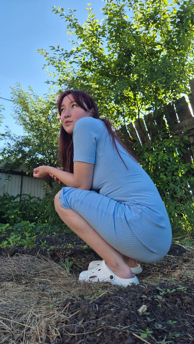 A young woman with long, dark hair and fair skin is crouching in a garden. She is wearing a light blue, ribbed, short-sleeved dress and white Crocs. The woman is looking upwards and to her left, with her right arm resting on her bent left knee. The ground in front of her is dark soil with some dried grass and small green plants. Behind her, a wooden fence can be seen, along with lush green trees and a bright blue sky. The setting appears to be a rural or suburban garden.