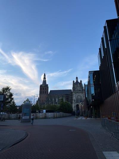 The image depicts a street scene in 's-Hertogenbosch, Netherlands, with the Sint-Janskathedraal (St. John's Cathedral) prominently featured. The cathedral, a Gothic structure, is partially covered in scaffolding, suggesting ongoing renovation or construction work. To the right of the cathedral, a modern building with dark metallic cladding and glass panels stands. The foreground shows a paved street with brickwork, a traffic island, and some temporary metal barriers. A single person is walking across the street towards the viewer. The sky is blue with wispy white clouds, and there appears to be a contrail from an airplane. The overall lighting suggests it is late afternoon or early evening. In the background, trees and some temporary fencing are visible.