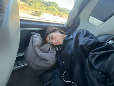 A young female is depicted reclining in a seat inside a passenger vehicle, identified as a bus by the 'Irizar' logo on a headrest, while traveling in the area of Santarém, Portugal. She possesses dark hair, light eyes, and a nose piercing. She is wearing a dark, long-sleeved button-up shirt over a dark top with a white graphic, and white wired earbuds are inserted into her ears with the cord visible. Her head rests on a light grey, bundled fabric item, possibly a garment or soft bag, positioned against the vehicle's interior side paneling, serving as a pillow. She looks directly towards the camera with a neutral expression. The vehicle's interior features dark and light grey seating. Through the window, a landscape of green, tree-covered hills under a bright, clear sky is visible, indicating an outdoor, possibly rural, setting in motion. A white gridded vent is visible near the floor of the vehicle.