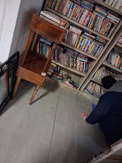 A tall bookshelf is filled with DVDs and books. A person kneels on the floor to the right of the shelf. An ornate wooden desk with a mirror leans against the bookcase, angled to the left. A metal frame lies on the floor near the desk. The room is illuminated by overhead lighting.