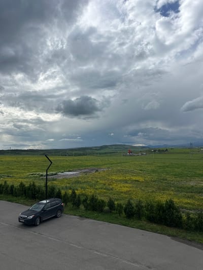 A black SUV is parked on an asphalt road adjacent to a green field. The road stretches into the distance, and the field is bordered by trees and vegetation. In the background, a landscape of rolling hills and mountains can be seen under a cloudy sky. The location is in the city of Marneuli, Georgia.
