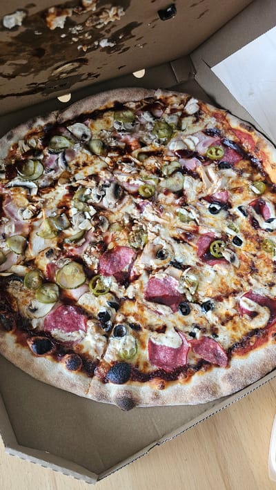 A close-up, top-down shot captures a pizza presented in a cardboard box. The pizza is generously topped with various ingredients, including melted cheese, sliced pepperoni, mushrooms, pickled cucumber slices, jalapeno pepper rings, black olives, and what appears to be pieces of chicken and possibly ham. A dark, glossy sauce, likely barbecue, is drizzled over the toppings. The pizza crust is golden brown with some charred edges, indicating it has been baked. The cardboard box is open, revealing its interior and the pizza within. The environment appears to be indoors, on a wooden surface, possibly a table. There are no people visible in the image. The scene does not suggest any specific location within the city of Slengiai, Lithuania; it depicts a generic food item in a common setting.