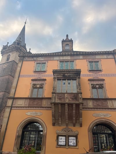 The image depicts the Hôtel de Ville (Town Hall) in Obernai, France, specifically focusing on its Renaissance-style facade and adjacent tower. The primary structure features an orange-hued facade with decorative stone elements. To the left, a slender, dark-stoned tower with a pointed spire rises, displaying a clock face (hands indiscernible) on its lower section.

The main facade is vertically stratified by horizontal decorative friezes in beige and pink/red tones. Multiple windows with stone frames and glass panes are visible across two upper levels; some are rectangular, others have pointed arched tops. Centrally positioned on the second level is a prominent, ornate oriel window, richly carved, with multiple glass panes, one of which shows a blue reflection or object. Above the main facade, an elaborate cornice leads to a pitched roof featuring a smaller, classical-style structure with an arched window.

On the ground floor, two large arched openings are fitted with black metal grilles and glass. Above the left opening, the word "ARCHIVES" is inscribed, and a planter with red flowers sits below the window. Above the right opening, "CORPS DE GARDE" is inscribed. Centered below the oriel window are two framed display boards exhibiting black and white images. The very bottom of the frame shows a dark, reflective surface, possibly part of a vehicle or street-level element. The sky above is overcast with diffuse clouds. No individuals are present in the scene.