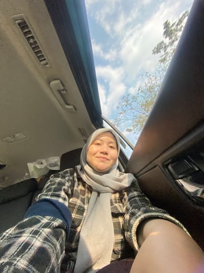 A woman wearing a light gray hijab and a black and white plaid shirt is inside a car. She is smiling and looking at the camera. The car has a sunroof open, revealing a blue sky with white clouds and some trees in the background. The interior of the car, including the ceiling with air vents and a handle, is visible. The scene appears to be in Mandar Sari, Indonesia, given the natural lighting and foliage.