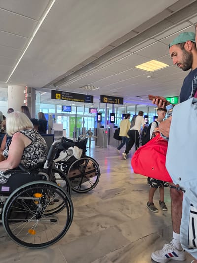 This image shows a group of people in what appears to be an airport or a similar transit hub, possibly in San Bartolomé, Spain. The scene is indoors, with bright overhead lighting and marble flooring. Several individuals are seen walking or standing, some of whom are in wheelchairs. One person in the foreground is holding a mobile phone, while another person in a light blue jacket is partially visible. Further down, a person in a red shirt and black shorts is also visible. Signs in English and Spanish are present, including "Alle Pass, All passports, Todos los pasaportes" and "Gate, Puerta 11". The presence of multiple gates and what seems to be a passport control area suggests this is an airport departure or arrival hall. The atmosphere appears to be busy, with people moving through the space, indicative of travel. The time of day is not explicitly stated, but the bright indoor lighting suggests it could be daytime. The overall impression is one of travel and transit.