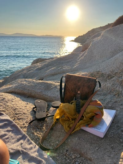 The scene depicts a rocky coastline at Agios Ioannis Diakoftis, Greece, under a clear sky with the sun low on the horizon, casting a bright reflection across the calm sea. Distant landmasses are visible on the horizon. In the foreground, several items are arranged on light-colored boulders: a grey towel partially covers a human leg, indicating the presence of an individual. Adjacent to the towel, two black flip-flops are positioned, one resting sole-down and the other inverted. To their right, a brown, hair-on-hide shoulder bag with a black strap stands upright. A crumpled yellow fabric, displaying faint orange and red stripes, rests in front of the bag, partially obscuring a dark, cylindrical object with a textured surface. To the right of the fabric, a white book with a visible spine and partial text, possibly "Writing Process," lies flat on the rock.