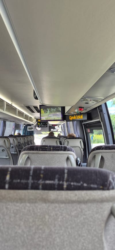 The image depicts the interior of a bus, likely in Norway, given the text "Gjøvik lokal" on a digital sign. The perspective is from a passenger looking down the aisle towards the front of the bus, with the image rotated 90 degrees counter-clockwise.

Several rows of grey fabric seats with dark patterned headrests and armrests are visible. Some seats have "VOLVO" printed on their headrests. Passengers are seated in some of the seats, their backs to the camera, with their hair visible. The overall impression is of a public transport vehicle.

In the foreground, on the left side of the bus, there is a large, light-colored panel that extends down the length of the visible interior. Attached to this panel is a screen displaying what appears to be landscape imagery, possibly a video or a static image. Above this screen, a yellow digital sign displays the text "Gjøvik lokal" in uppercase letters. Another smaller screen or sign is visible further down the aisle.

The scene suggests a daytime journey. The lighting seems natural, with some ambient light coming from the windows, which are partially visible on the right side of the image, showing greenery outside. The mood is calm and ordinary, as one might expect on a bus during transit. There are no strong emotional cues evident. The setting is clearly the interior of a passenger bus.