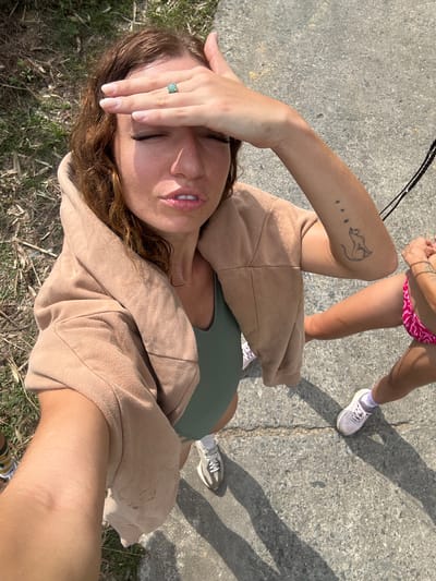 A selfie, angled downward, shows a woman's face with eyes closed and a hand on her forehead wearing a turquoise ring. She has brown hair, fair skin, and is wearing a green top and a tan hooded jacket. The background includes a concrete surface, a second person wearing a pink patterned bathing suit bottom, and partial views of shoes. There is also grass and shadows visible. The location is likely outdoors, with a mix of pavement and grassy areas. It is unlikely to be Suối Giàng, Vietnam, given that bathing suits are not commonly worn in that area of Vietnam.