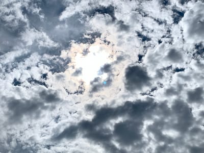The image shows a sky filled with clouds, with the sun partially obscured by them. The clouds range in color from bright white to dark gray, indicating varying degrees of density and thickness. The sun is visible in the center of the frame, creating a bright halo effect as its light diffuses through the cloud cover. There are no people, structures, or objects visible in this image, only the sky and its cloud formations. The location in Smorodikha, Russia is not discernible from the image itself.