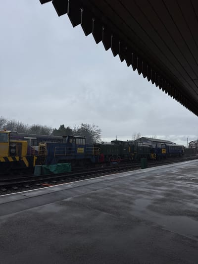 The scene depicts a railway station platform and tracks under an overcast sky in Cannock Chase, United Kingdom. The dark grey asphalt platform is visibly wet with numerous puddles, indicating recent precipitation. White lines delineate the platform edge, adjacent to which an orange cable runs between the railway sleepers. A dark green rectangular bin stands on the platform near the tracks.
On the adjacent railway tracks, a line of stationary rolling stock and locomotives is arranged. From left to right, a yellow and black locomotive or shunter is partially visible. Behind it, a purple passenger carriage is present. Next is a blue locomotive, marked with the number "5" and the text "N.C.B. OPENCAST EXECUTIVE" on its side, featuring yellow handrails. A green tarp lies on the ground beside this locomotive. Following this are two dark green locomotives; the first has prominent red wheels. Further along the track, a blue and yellow striped unit precedes a blue passenger carriage labeled "M&CR LADY CHESWOOD," followed by additional blue carriages extending out of view to the right.
In the background, a dense line of bare deciduous trees is visible on the left. To the right, behind the parked trains, a single-story grey building with a dark roof is observed. Further right, a small signal box or utility building with a red roof is discernible. The sky is uniformly grey and overcast. No individuals are present within the captured frame, and no active movement is apparent.