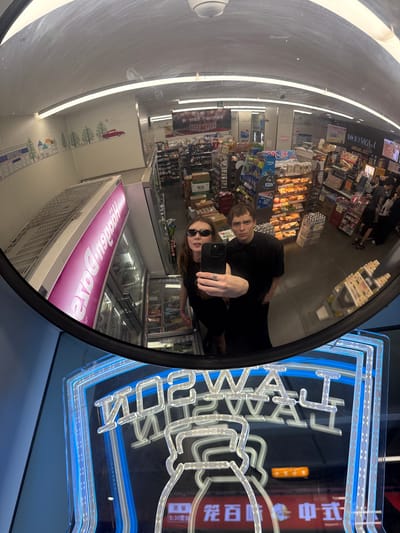 The image is a mirror selfie featuring two individuals inside a Lawson convenience store located in Changning District, China. A woman with long brown hair, wearing dark sunglasses and a dark top, stands on the left, holding a dark-colored smartphone with a silver ring visible on her left hand, taking the photograph. Beside her, a man with short brown hair, also dressed in a dark shirt, looks towards the camera.
The mirror's reflection reveals the interior of the convenience store: aisles are stocked with various packaged goods, a prominent display case holds baked goods/pastries, and multiple refrigerated units are visible. A large freezer unit on the left side of the reflection prominently displays the upside-down and reversed "Häagen-Dazs" brand name in pink. An upper wall features a cartoonish mural depicting small houses, trees, and a red car, alongside the word "LAWSON" written in reverse. Overhead, fluorescent tube lighting illuminates the store, and a ceiling-mounted security camera is discernible. At the bottom of the mirror, a blue and white neon sign with the reversed "LAWSON" logo is visible. Various signs and product packaging throughout the store display Chinese characters. Other customers are faintly visible in the deeper background aisles.