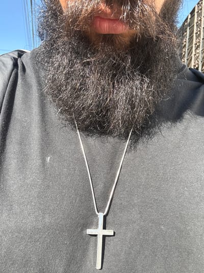 A close-up, ground-level shot shows a person with a dense, graying beard wearing a black shirt and a silver cross necklace. Only the lower portion of the face is visible. The background is blurred, but appears to be an urban setting. The location is not discernible from the given image.